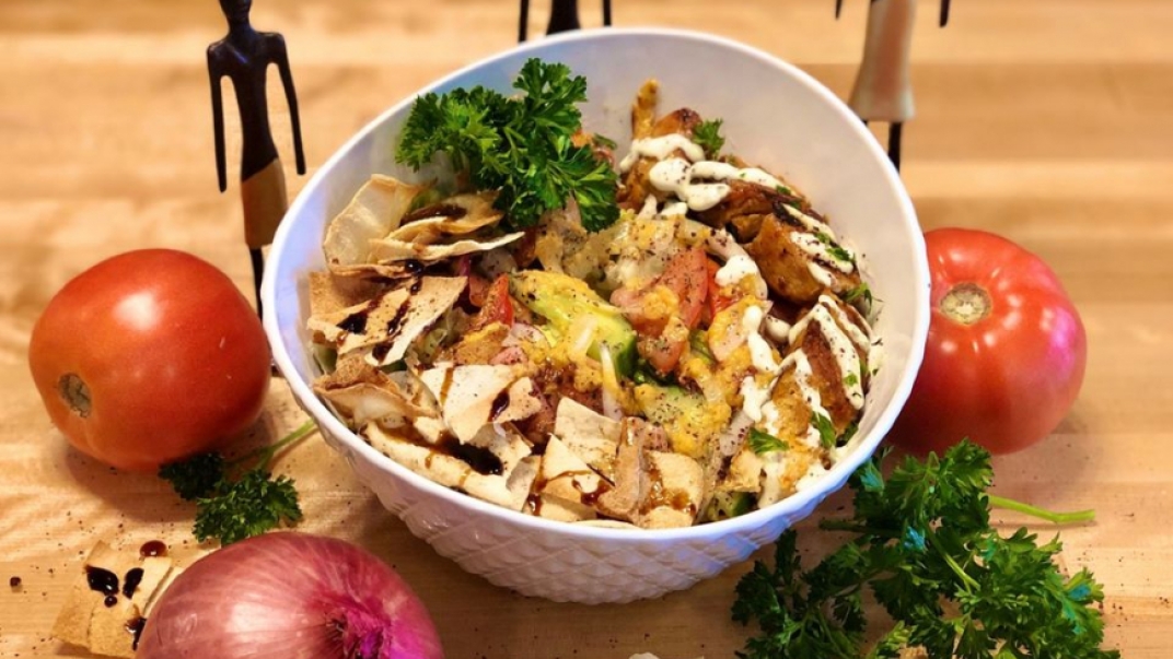 A bowl of food with pita chips crumbled into the bowl, cilantro, and meat from a kebab drizzled with sauce. There are whole tomatoes and an onion around the bowl. 